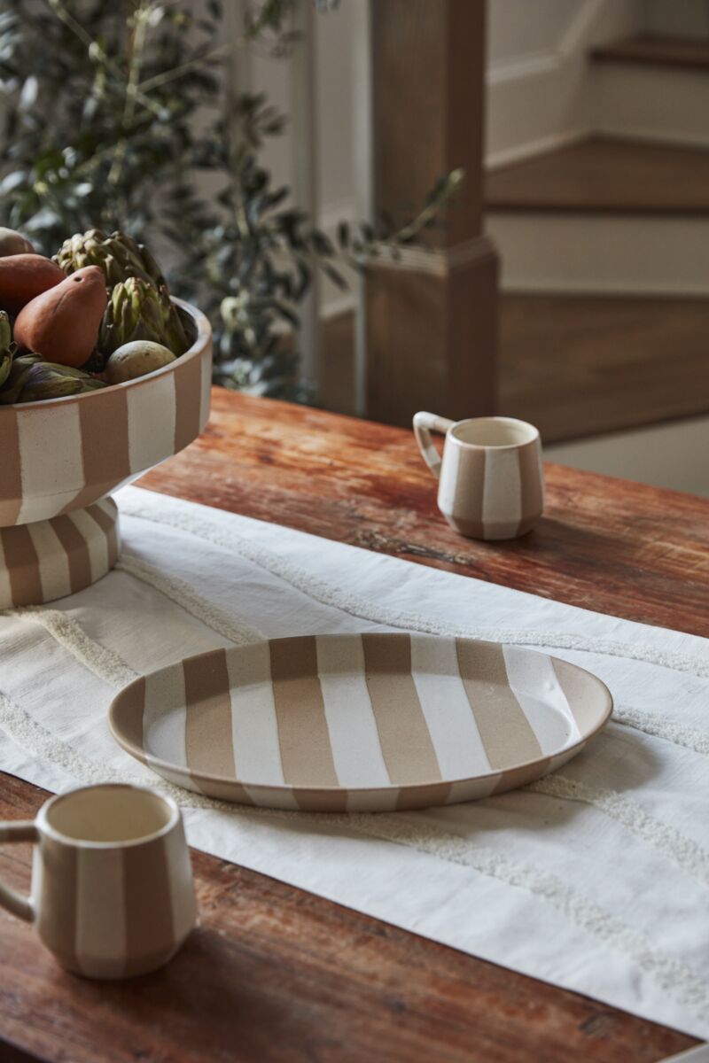 A rustic wooden table features minimalist dinnerware: the beige and white striped Botera Platter by Accent Decor, two matching mugs, and a large fruit bowl on a white runner, with a staircase and leafy plant in the background.