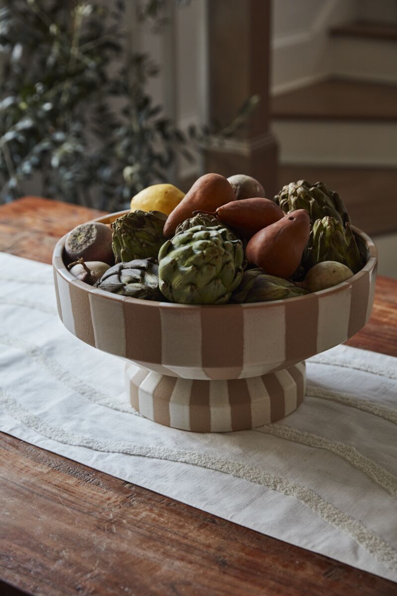 A beige and white striped Botera Footed Bowl by Accent Decor sits on a wooden table filled with artichokes, pears, and potatoes. In the background are a blurred staircase and leafy plant. This ceramic serving dish adds style to any setting.