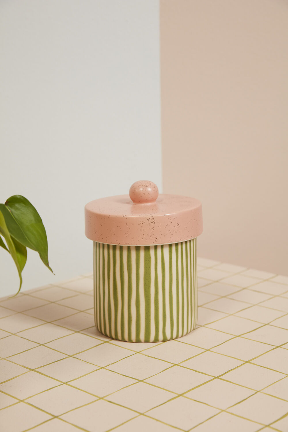 The Margot Canister by Accent Decor, featuring hand-painted green and white vertical stripes and a round pink lid, rests on a grid-tiled surface with a green plant leaf visible to the left.