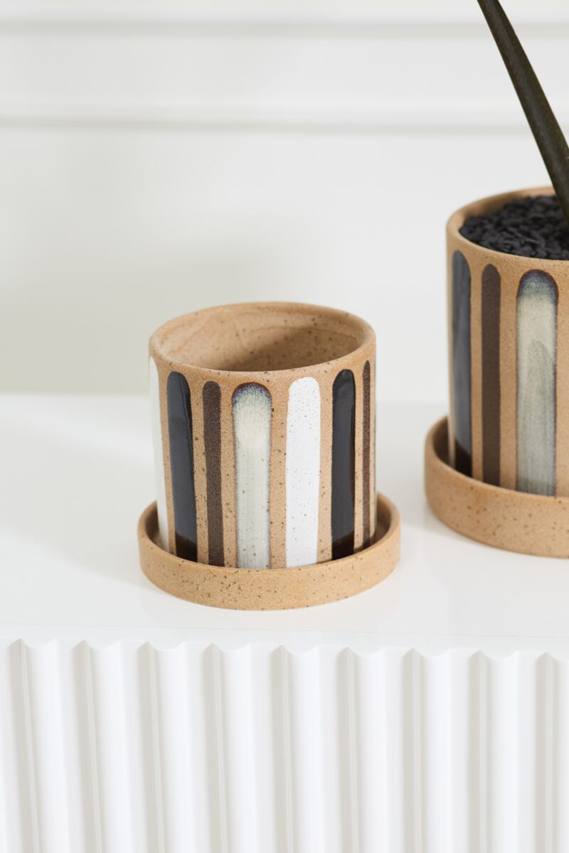 The Truman Pot/Saucer 6x5.5" by Accent Decor, a small round hand-painted ceramic pot with a tan base and vertical black, gray, and white stripes, sits beside a larger matching planter on a textured white surface.