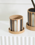 The Truman Pot/Saucer 6x5.5" by Accent Decor, a small round hand-painted ceramic pot with a tan base and vertical black, gray, and white stripes, sits beside a larger matching planter on a textured white surface.