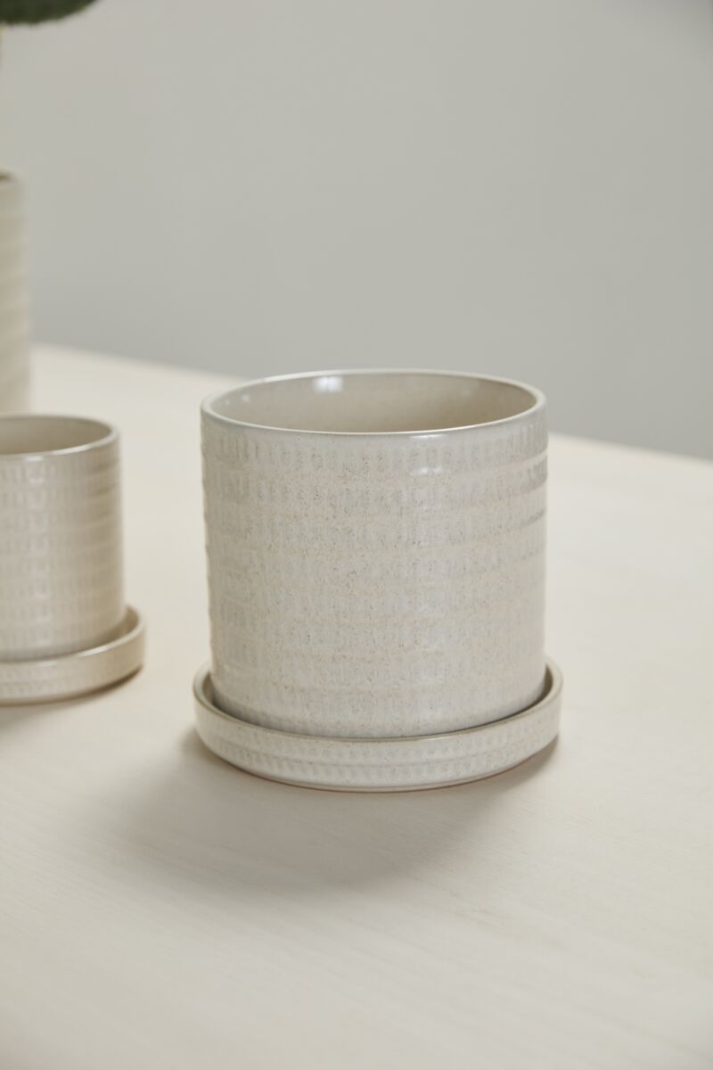 An Accent Decor Morse Pot /Saucer in cream with a drainage hole rests on a light-colored table, while another similar pot is partially visible in the background.