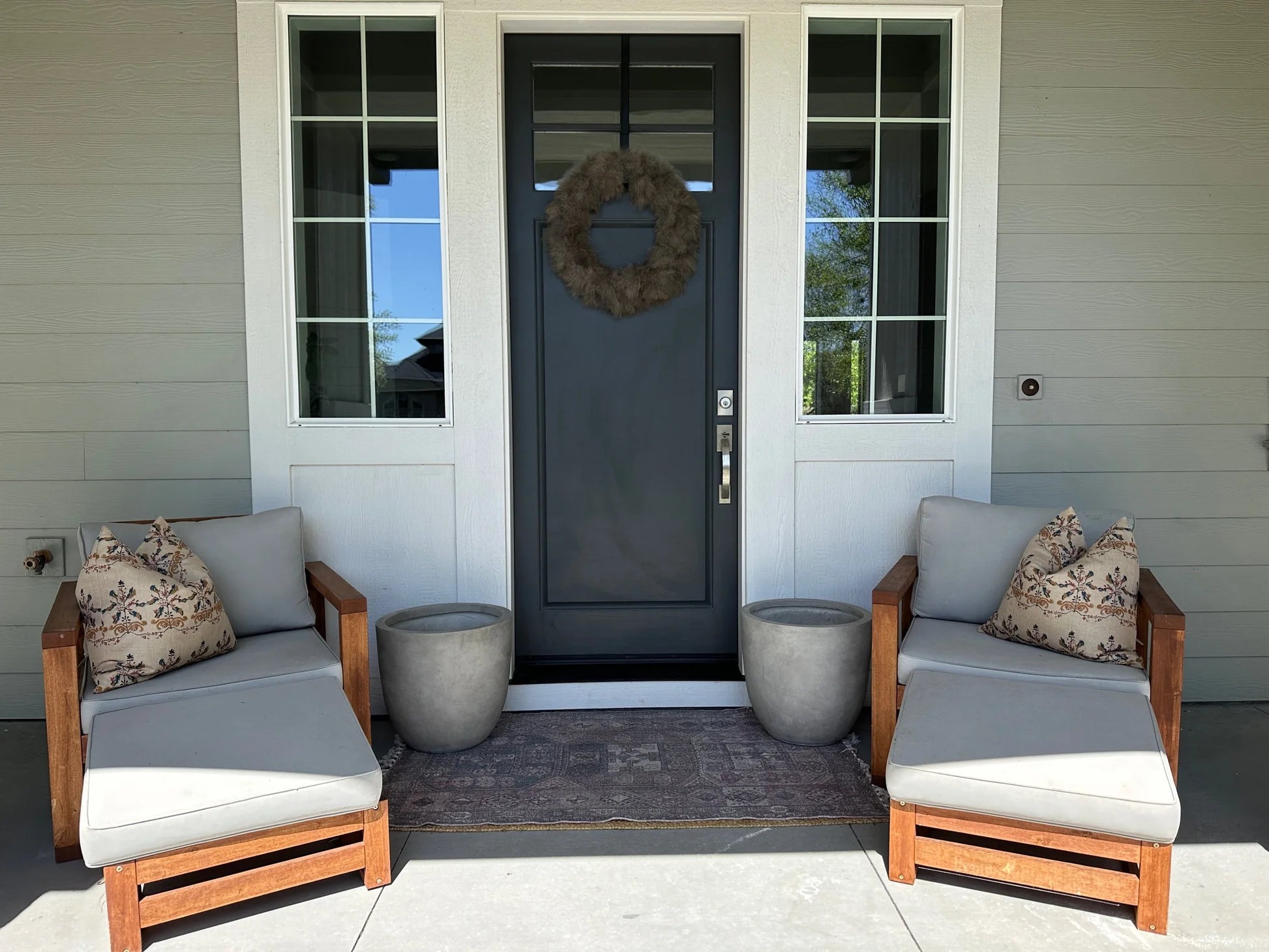 A front porch with a dark door, wreath, two cushioned wooden chairs featuring Rennah Collective Birdie Pillow 22x22 covers, two empty gray planters, and a small rug on a concrete floor.