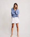 A woman poses against a plain background wearing Denimist's HAISLEY SHORT WHITE 100% cotton frayed shorts, a blue and white striped button-up shirt, and white sandals. She has shoulder-length hair and touches her face in a relaxed stance.