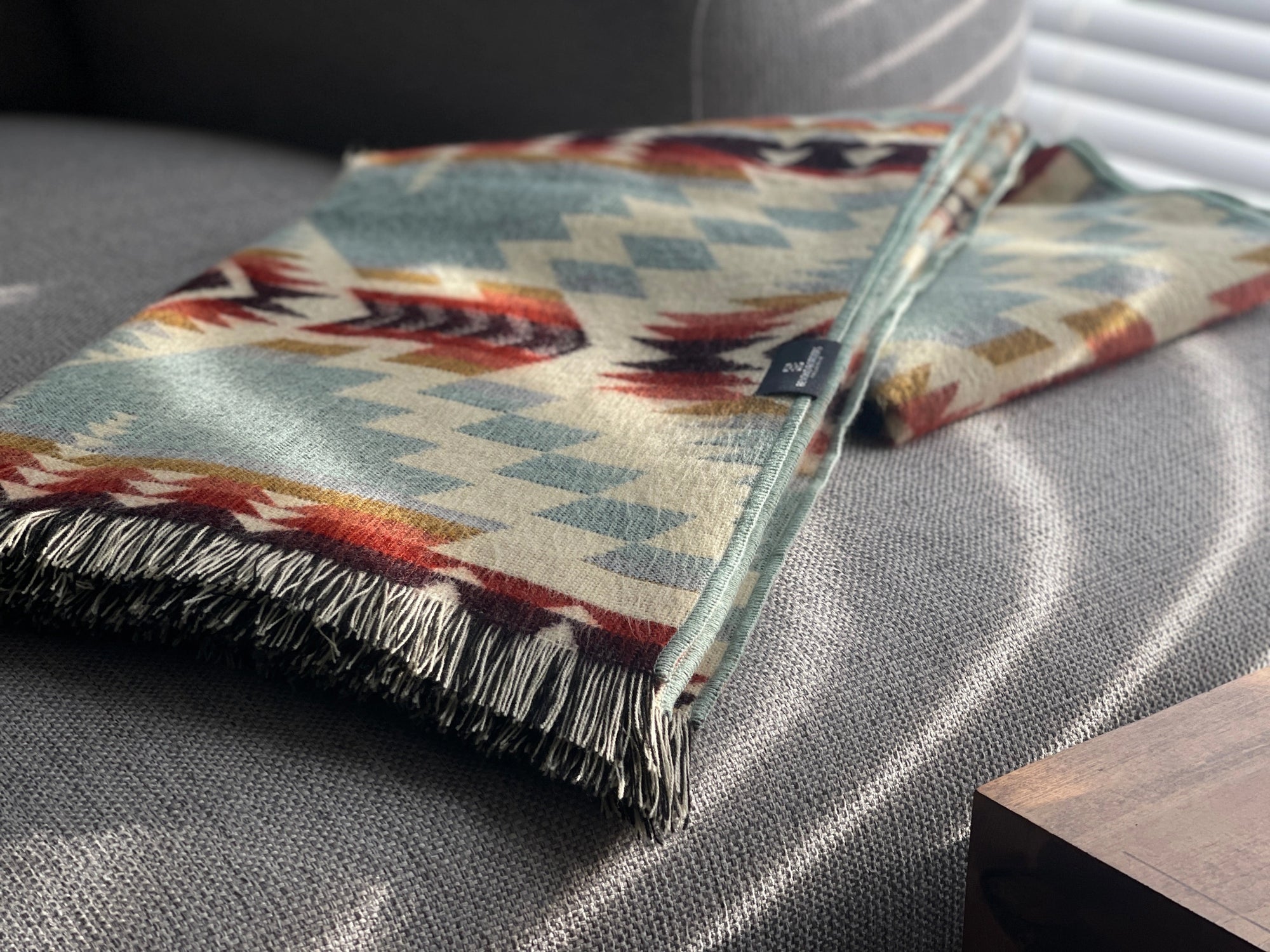 A folded Secoya Throw by Faire, featuring a geometric pattern in teal, cream, red, and brown and crafted by Indigenous Ecuadorian artisans, rests on a gray couch lit by sunlight filtering through blinds.