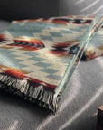 A folded Secoya Throw by Faire, featuring a geometric pattern in teal, cream, red, and brown and crafted by Indigenous Ecuadorian artisans, rests on a gray couch lit by sunlight filtering through blinds.