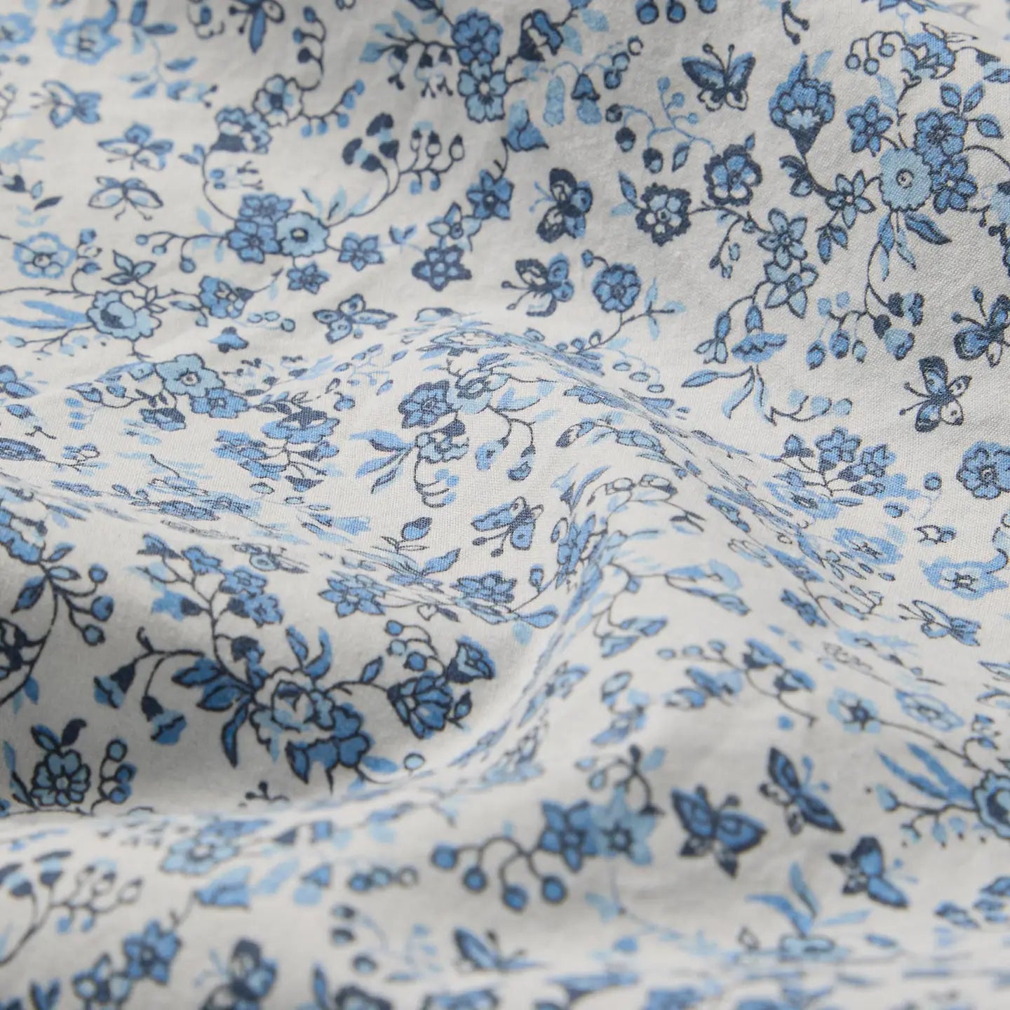 A close-up of Faire’s Blue Mini Meadow Euro Sham shows 100% cotton percale with a white background and small blue floral pattern, featuring gentle folds and shadows from the slightly wrinkled fabric.