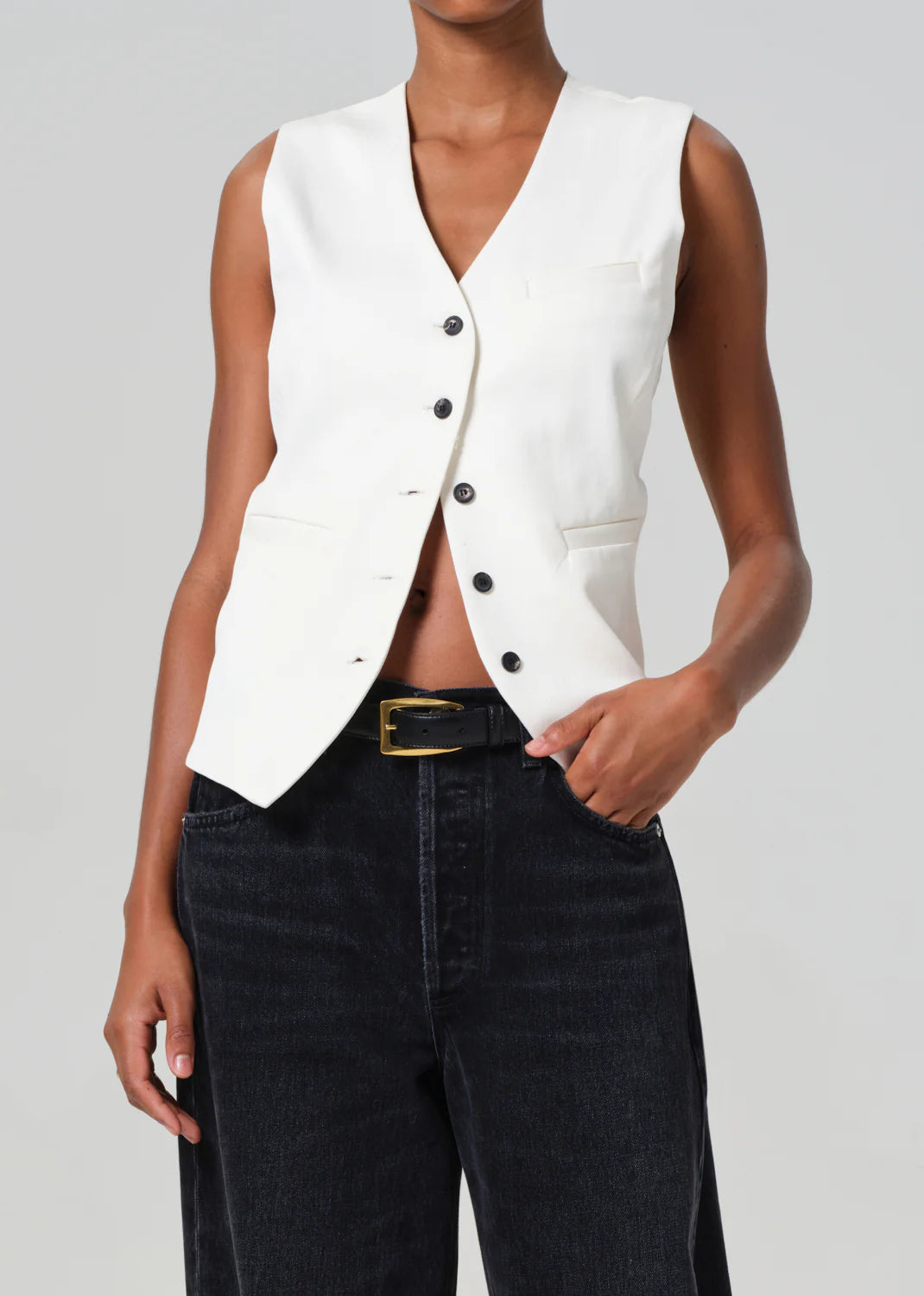 A person models the Jasmin Long Vest by Citizens Of Humanity/AGOLDE, made from regenerative cotton. The white vest with black buttons and pockets enhances the tonal look against a neutral background, paired elegantly with sleek black jeans.