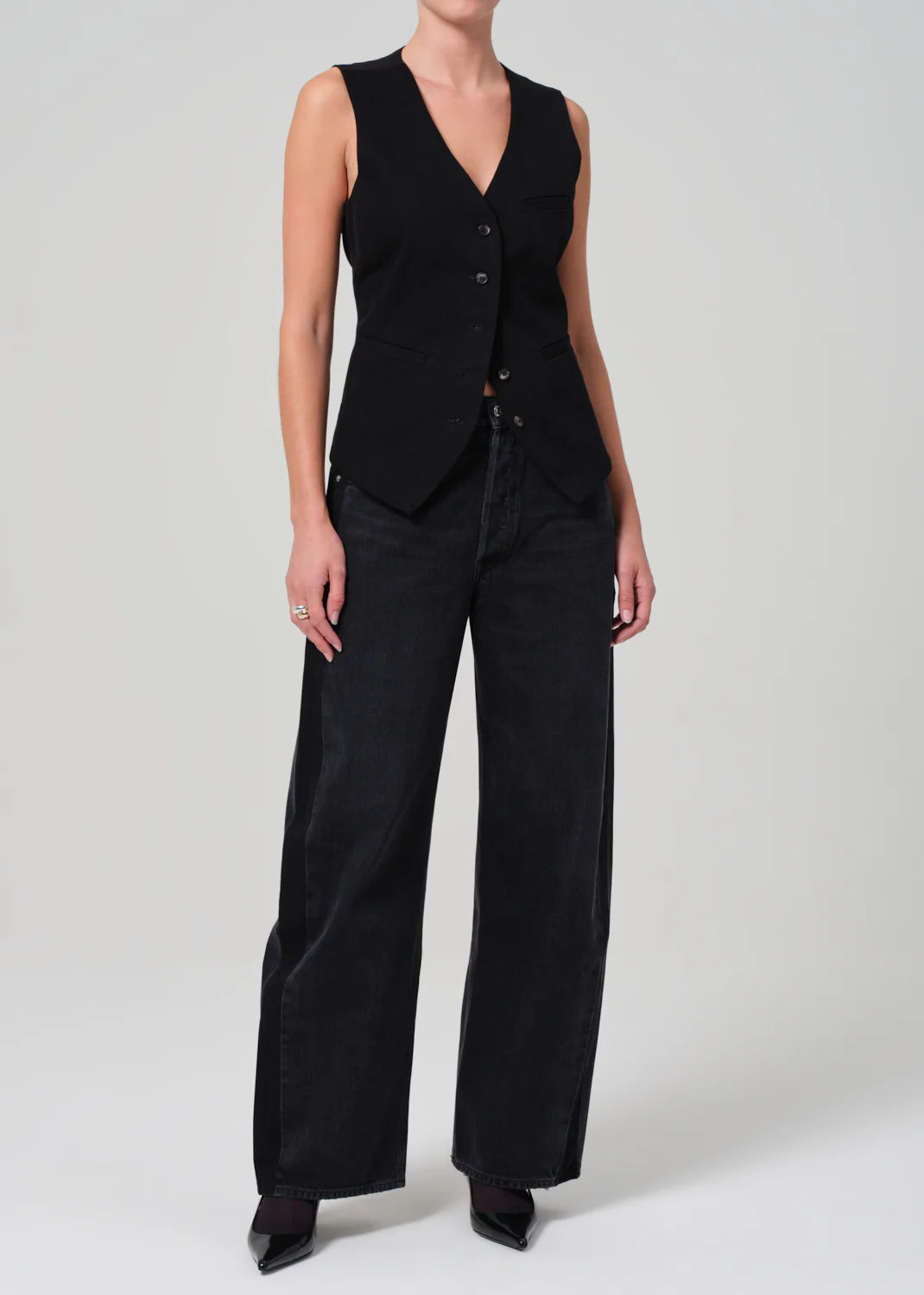 A person wearing a Jasmin Long Vest from Citizens Of Humanity/AGOLDE, paired with wide-legged black jeans and pointed black shoes, elegantly exemplifies tonal dressing against a simple backdrop.