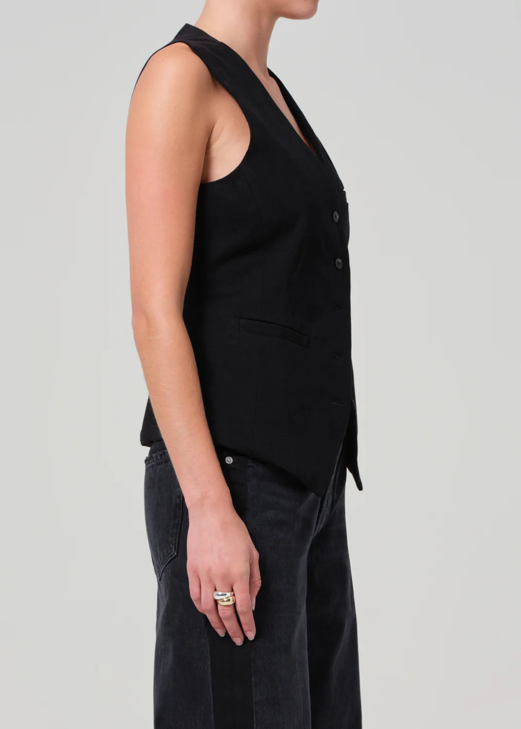 A person wears a Jasmin Long Vest from Citizens Of Humanity/AGOLDE, paired with black jeans, standing in profile against a plain backdrop. With their hands at their sides, they display rings on their fingers, embracing tonal dressing in a sleek ensemble.