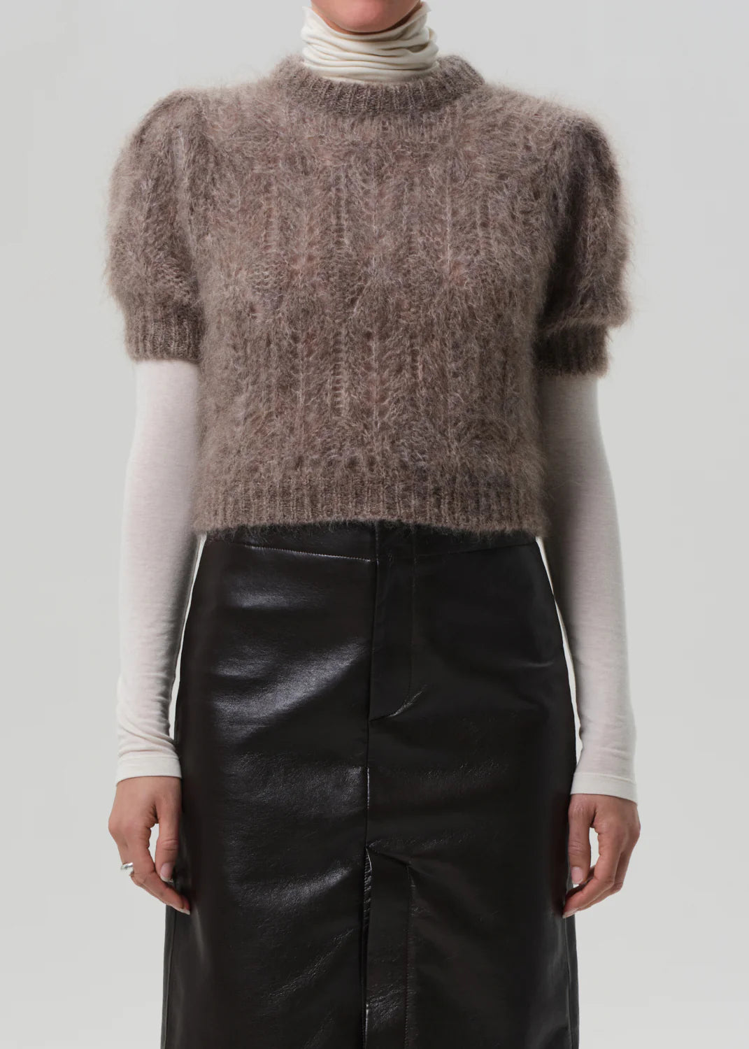 Someone wears the odette puff sleeve sweater by Citizens Of Humanity/AGOLDE, layered over a long-sleeved cream turtleneck and paired with a high-waisted black leather skirt, set against a plain light background.