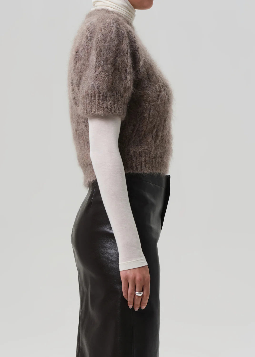 A person stands in profile against a plain light gray background, wearing the Citizens Of Humanity/AGOLDE odette puff sleeve sweater in taupe over a white turtleneck and a high-waisted black leather skirt. Their ring-adorned left hand rests by their side.