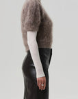 A person stands in profile against a plain light gray background, wearing the Citizens Of Humanity/AGOLDE odette puff sleeve sweater in taupe over a white turtleneck and a high-waisted black leather skirt. Their ring-adorned left hand rests by their side.