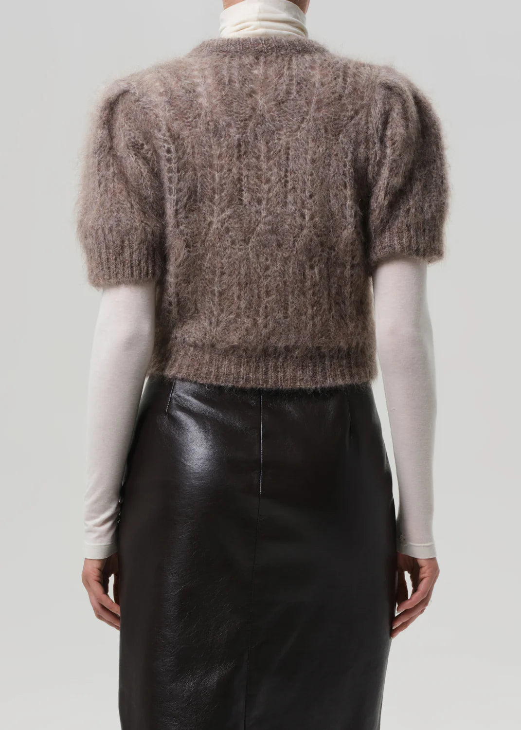 A person stands facing away, wearing the Citizens Of Humanity/AGOLDE odette puff sleeve sweater over a white turtleneck and a high-waisted black leather skirt against a plain, light background.