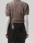 A person stands facing away, wearing the Citizens Of Humanity/AGOLDE odette puff sleeve sweater over a white turtleneck and a high-waisted black leather skirt against a plain, light background.