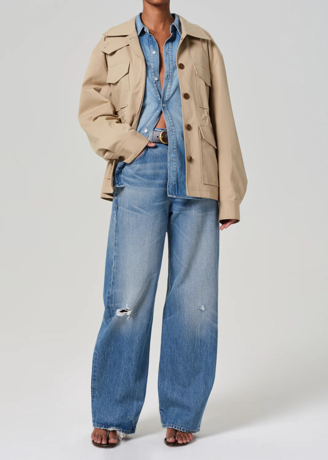 The individual wears a Paretti Military Jacket from Citizens Of Humanity/AGOLDE over a blue denim shirt, paired with wide-leg jeans featuring slight distressing and a dark belt. They are barefoot against a plain background.