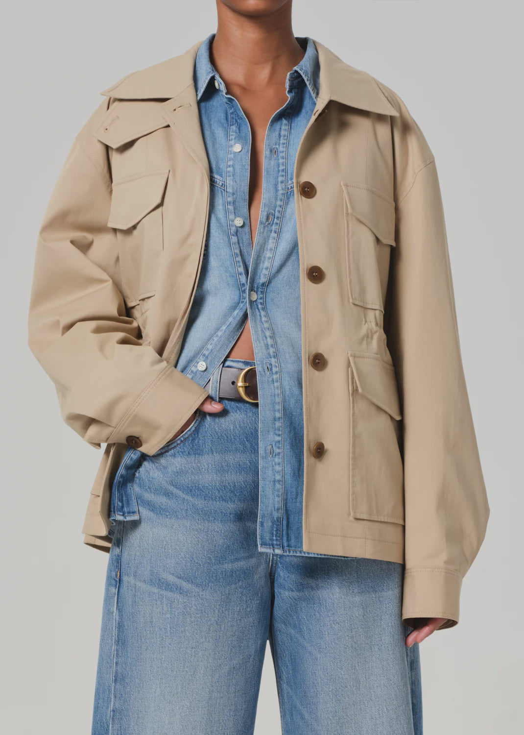 A person is wearing the Paretti Military Jacket from Citizens Of Humanity/AGOLDE, a tan utility style with front pockets and brown buttons, over a partially buttoned denim shirt and wide-leg denim jeans, accented by a brown belt. The background is plain light gray.