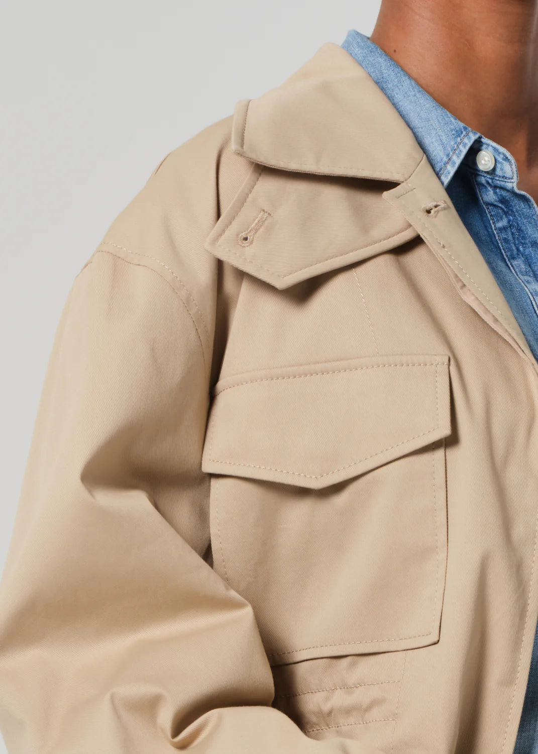 Someone wearing the Paretti Military Jacket by Citizens Of Humanity/AGOLDE in light brown cotton with large front pockets over a denim shirt. The image focuses on the jacket's collar and upper part against a light gray background, with the person's face not visible.