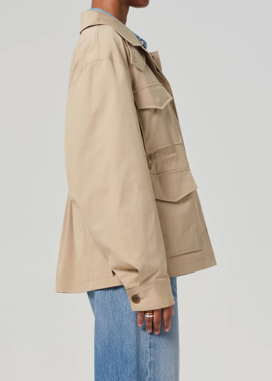 A person wearing the Paretti Military Jacket by Citizens Of Humanity/AGOLDE, a beige utility design with a drawstring waist, multiple large pockets, and a relaxed fit, is shown from the side against a plain background with blue jeans.