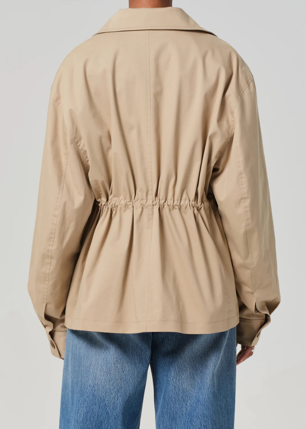 Rear view of a person in the Paretti Military Jacket by Citizens Of Humanity/AGOLDE, featuring a beige cotton design with a loose fit, drawstring cinched waist, and long sleeves paired with blue jeans against a neutral background.