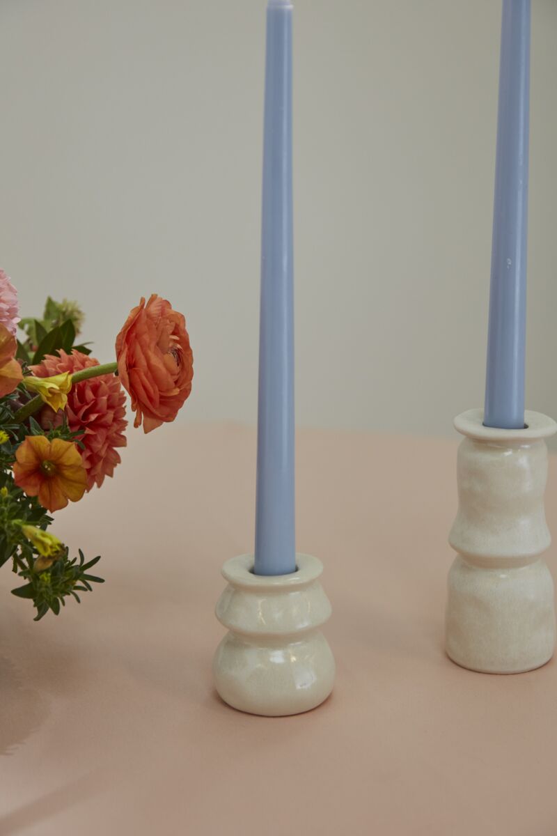 A bouquet of orange and pink flowers sits beside two light blue taper candles in Accent Decor’s Lyam Candleholders, featuring a white reactive glaze, on a pale pink surface for charming handmade decor against a neutral background.