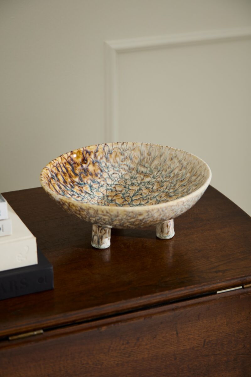 The Accent Decor Walden Bowl, a shallow, speckled ceramic piece with three short legs, sits on a dark wooden surface near books and a light-colored wall—ideal as sculptural home decor.