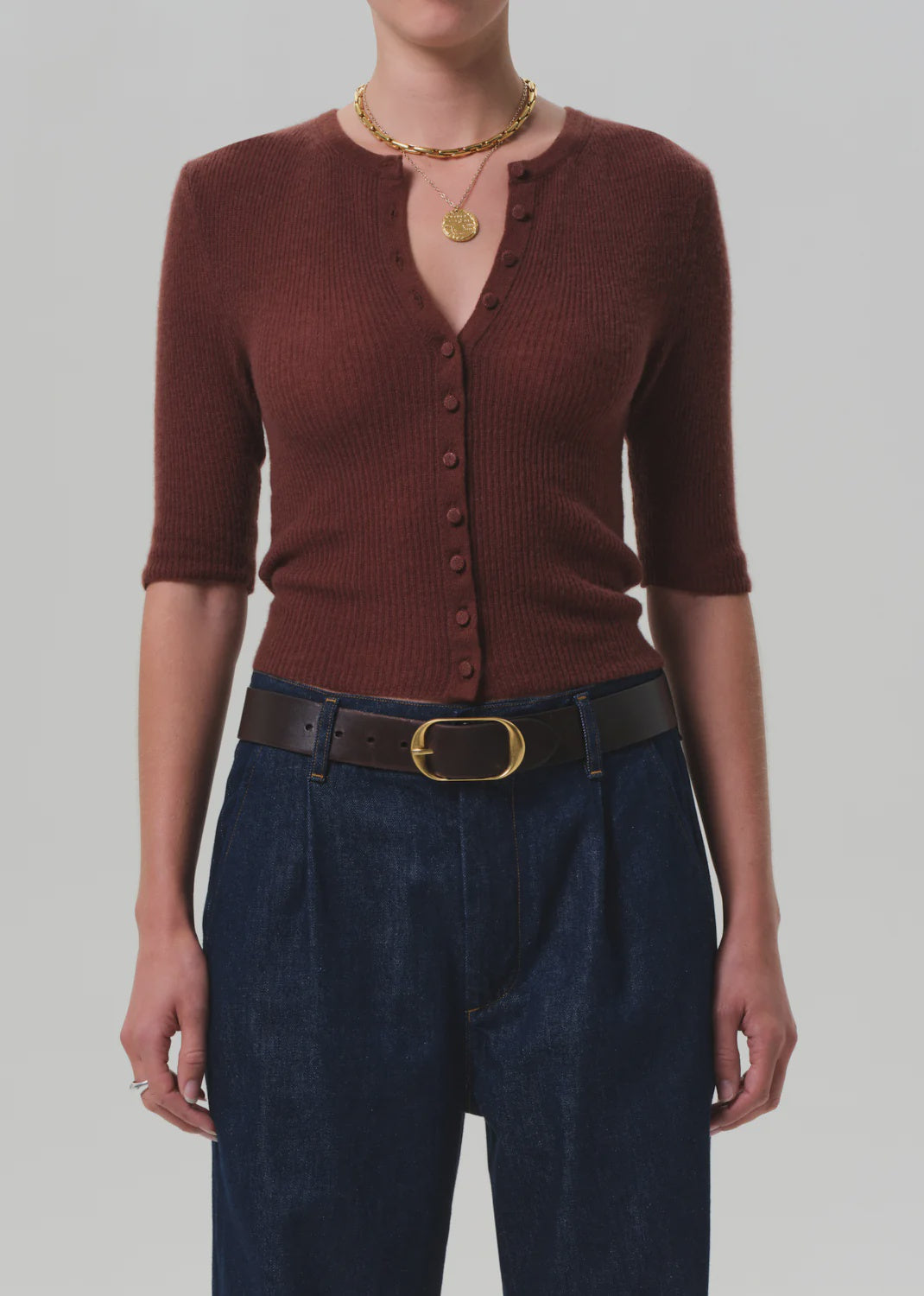 A person wearing the Tabitha Cardigan by Citizens Of Humanity/AGOLDE—slim-fit, ribbed, brown, with elbow-length sleeves—paired with dark blue high-waisted jeans, a brown belt with a gold buckle, and layered gold necklaces.