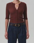 A person wearing the Tabitha Cardigan by Citizens Of Humanity/AGOLDE—slim-fit, ribbed, brown, with elbow-length sleeves—paired with dark blue high-waisted jeans, a brown belt with a gold buckle, and layered gold necklaces.