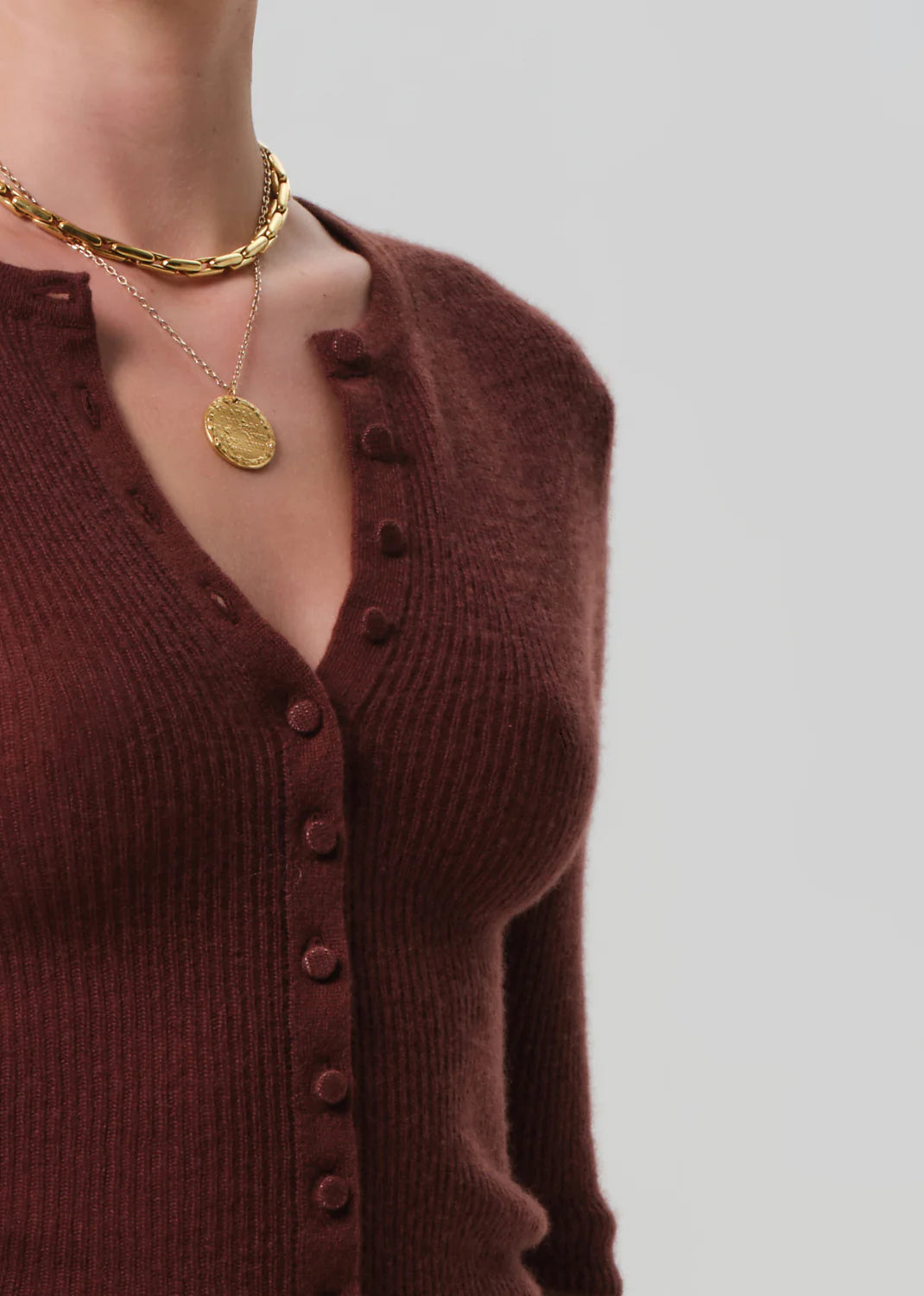 A person wearing the Tabitha Cardigan by Citizens Of Humanity/AGOLDE, a slim-fit maroon wool-cashmere blend with a deep V-neck and button-up front, styled with layered gold necklaces, poses against a light gray background.