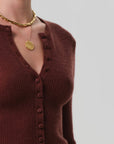 A person wearing the Tabitha Cardigan by Citizens Of Humanity/AGOLDE, a slim-fit maroon wool-cashmere blend with a deep V-neck and button-up front, styled with layered gold necklaces, poses against a light gray background.