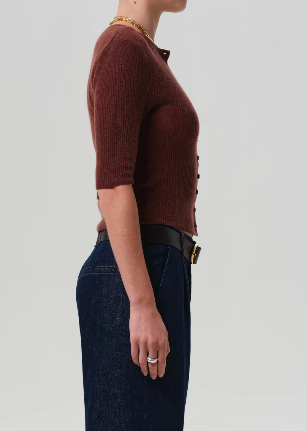 A person in profile against a light gray background wears the Tabitha Cardigan by Citizens Of Humanity/AGOLDE, paired with dark blue high-waisted jeans, a black belt, a gold necklace, and silver rings.