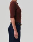 A person in profile against a light gray background wears the Tabitha Cardigan by Citizens Of Humanity/AGOLDE, paired with dark blue high-waisted jeans, a black belt, a gold necklace, and silver rings.