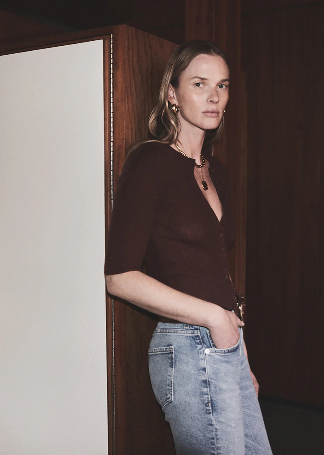 A woman with long hair stands indoors against a wooden wall, wearing the Tabitha Cardigan by Citizens Of Humanity/AGOLDE, paired with light blue jeans and gold jewelry, looking toward the camera with a neutral expression.
