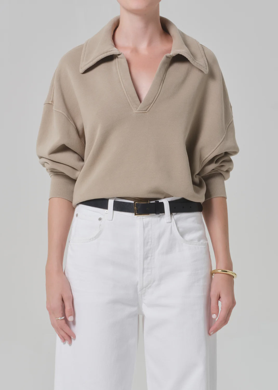 A person wears a loose taupe Citizens Of Humanity/AGOLDE trapeze pullover tucked into high-waisted white jeans, accessorized with a black belt and gold bracelet, standing against a neutral background.