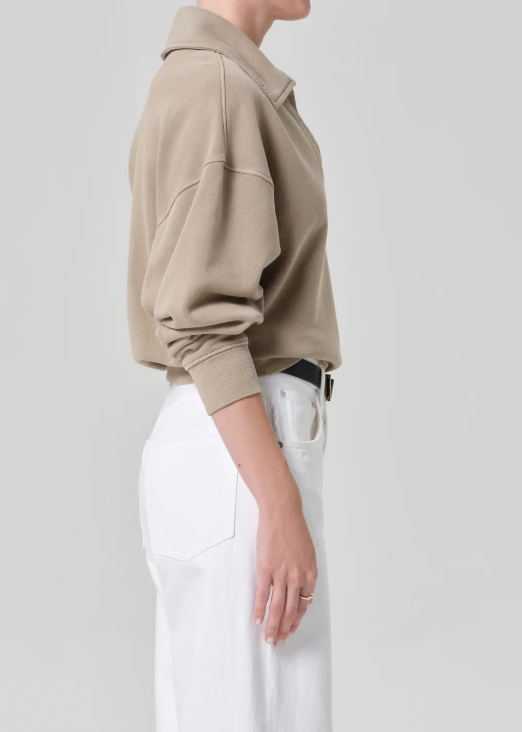 A person in profile wears the Citizens Of Humanity/AGOLDE trapeze pullover in beige sueded fleece, tucked into high-waisted white jeans with a black belt and a simple ring, set against a plain light gray background.