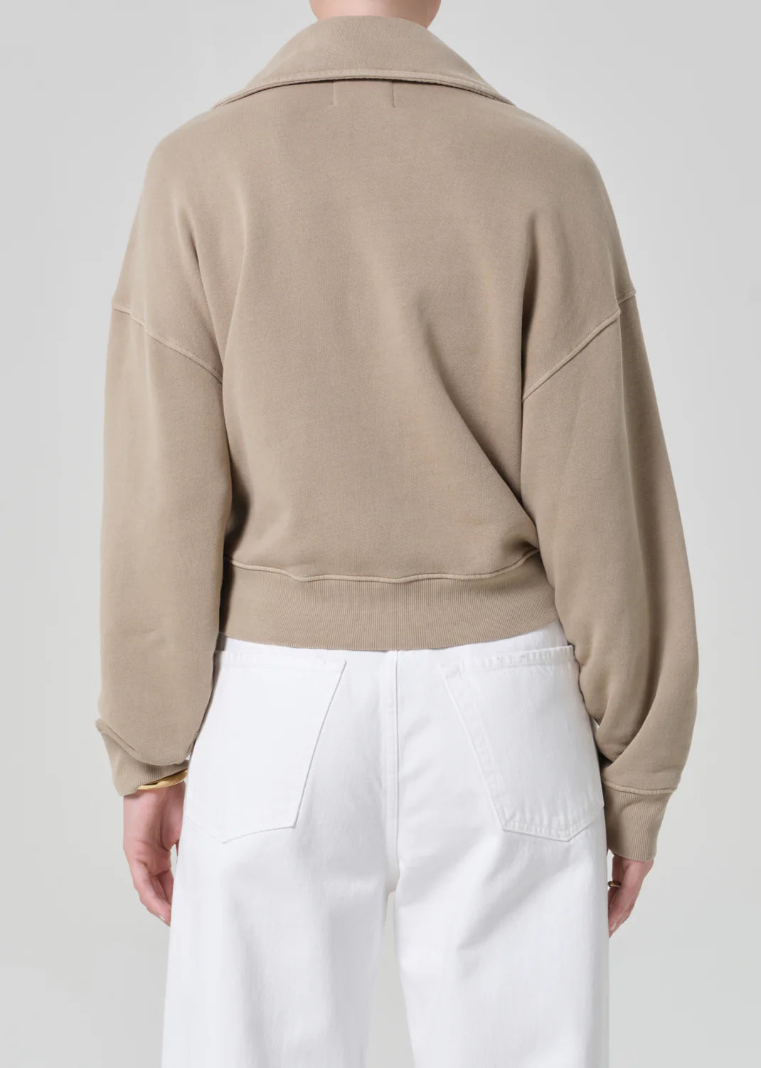 A person is shown from the back, wearing a light brown Citizens Of Humanity/AGOLDE trapeze pullover with a collar and high-waisted white pants against a plain, light gray background.