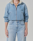 A model wearing the Citizens Of Humanity/AGOLDE bubble bomber in marseille stands against a light gray background, paired with high-waisted jeans and accessorized with a gold necklace and rings.