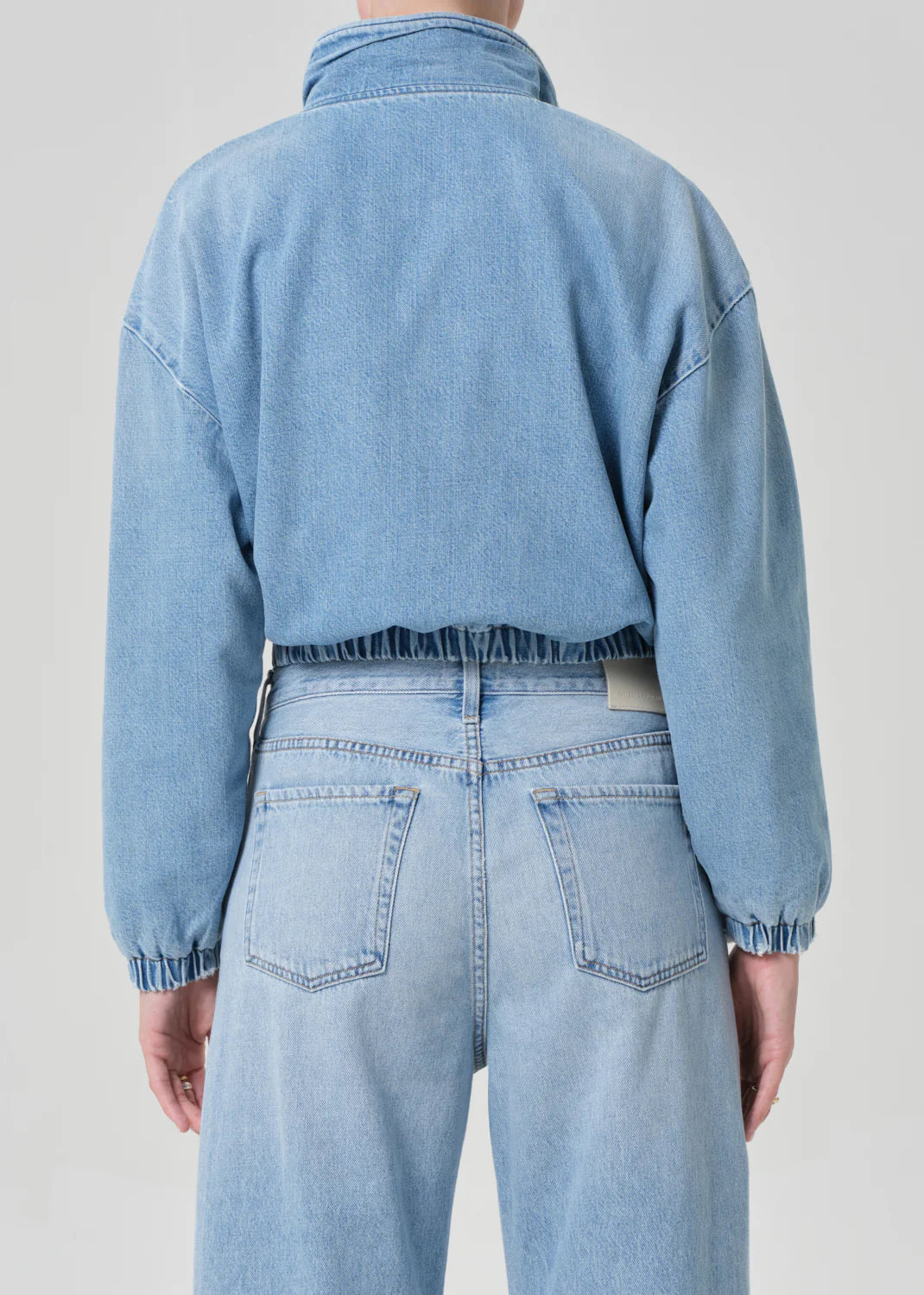 A person is shown from the back wearing the Citizens Of Humanity/AGOLDE bubble bomber in marseille, a light blue denim jacket with elastic cuffs and waistband, paired with matching high-waisted light blue vintage indigo wash jeans.