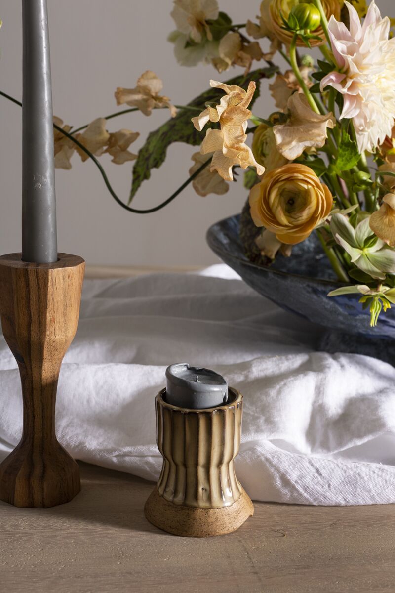 A close-up of a table featuring the Accent Decor Argon Candleholder in brown holding a gray candle, alongside a ceramic candleholder with a melted candle and a floral arrangement in a blue vase on white cloth.