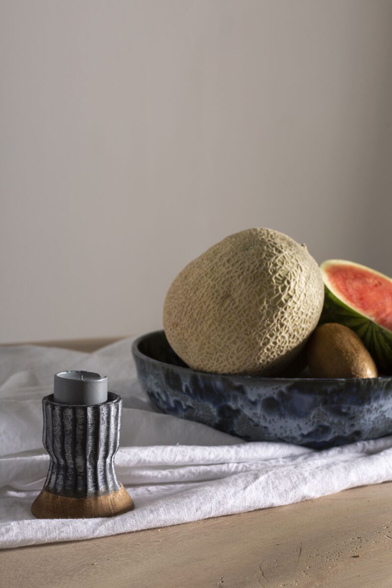 A ceramic bowl with a cantaloupe, watermelon slice, and kiwi sits on a white cloth beside the Accent Decor 51895 Argon Candleholder in blue, displayed on a wooden surface.