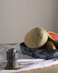 A ceramic bowl with a cantaloupe, watermelon slice, and kiwi sits on a white cloth beside the Accent Decor 51895 Argon Candleholder in blue, displayed on a wooden surface.