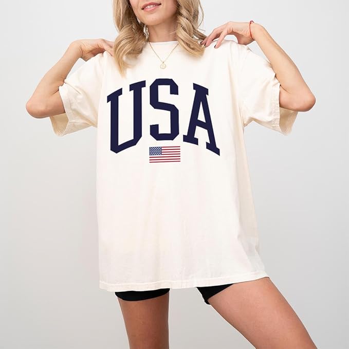 A woman wears the Faire USA Flag Tee—an oversized cream cotton shirt with bold navy "USA" lettering and a small flag, paired with black shorts. Made from soft-touch fabric for comfort, styled on a plain white background.