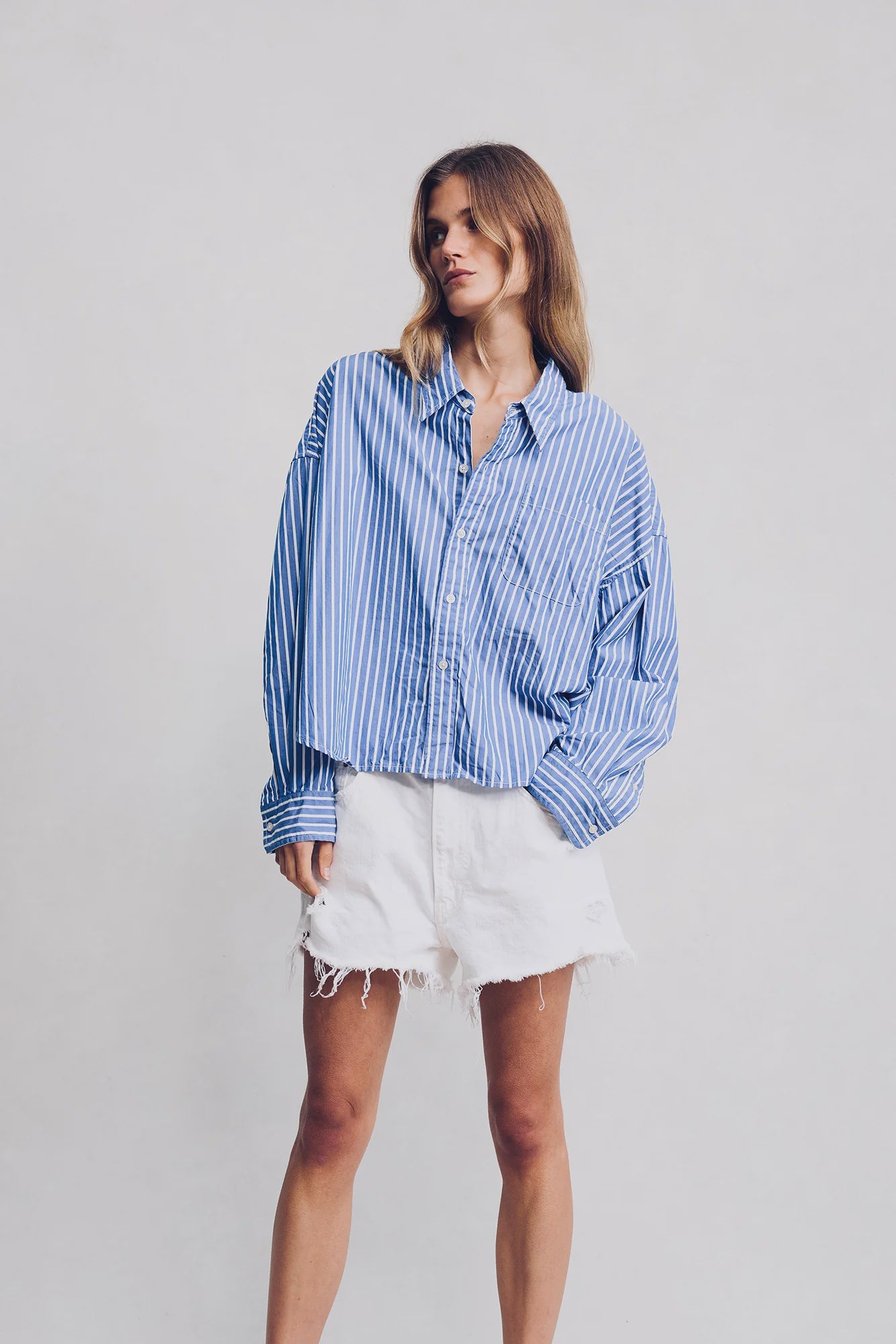 A woman in Denimist's HAISLEY SHORT WHITE and a loose blue-and-white striped button-up shirt stands against a plain light background, posing relaxedly and gazing to the side.