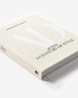 Architectural Digest at 100" by Hachette Book Group is a hardcover book celebrating Architectural Digest’s design legacy, featuring a light beige cover with embossed white "AD" letters and a ribbon bookmark, shown on a white background.
