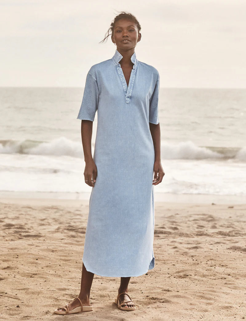 A woman stands barefoot on a sandy beach wearing the Emma Popover Henley Dress by Frank &amp; Eileen, featuring light blue fabric, short sleeves, a ruffled collar, and buttons. Ocean waves and a cloudy sky create a serene backdrop.