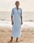 A woman stands barefoot on a sandy beach wearing the Emma Popover Henley Dress by Frank & Eileen, featuring light blue fabric, short sleeves, a ruffled collar, and buttons. Ocean waves and a cloudy sky create a serene backdrop.
