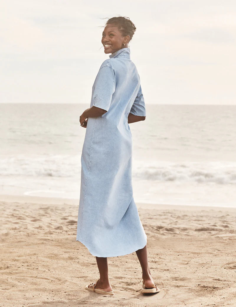 A woman smiles barefoot on a sandy beach, wearing the Frank &amp; Eileen Emma Popover Henley Dress in light blue, with the ocean and sky behind her.
