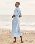 A woman smiles barefoot on a sandy beach, wearing the Frank & Eileen Emma Popover Henley Dress in light blue, with the ocean and sky behind her.
