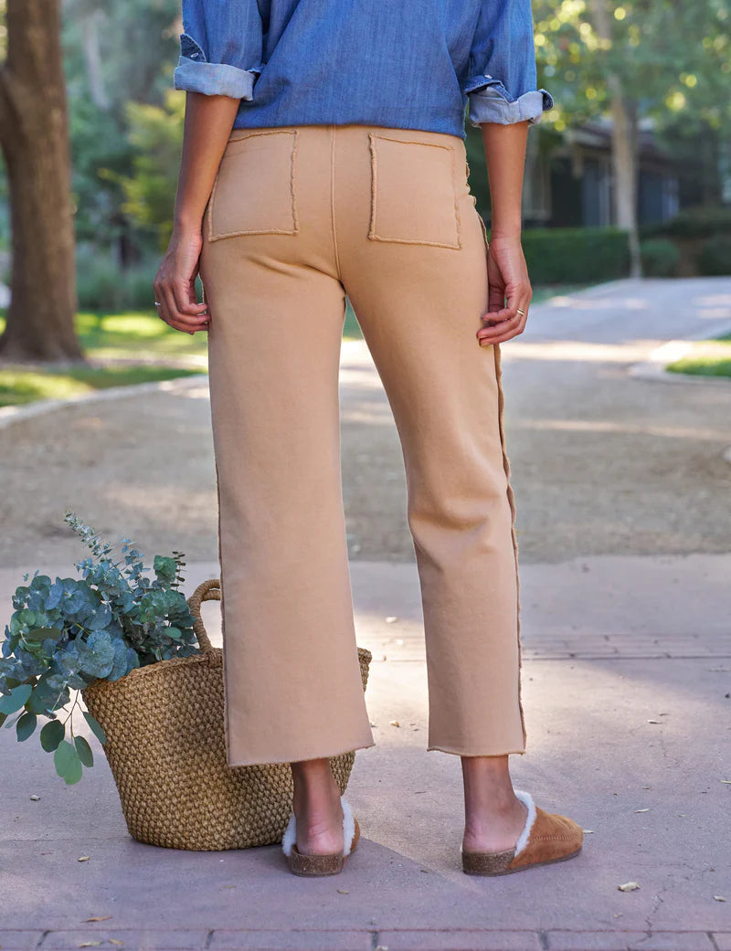 Outdoors on a path, a person faces away wearing a rolled-up denim shirt and Frank & Eileen’s Bella Sweatpant—100% cotton, wide-leg with patch pockets—paired with tan slip-on shoes beside a woven basket of greenery.