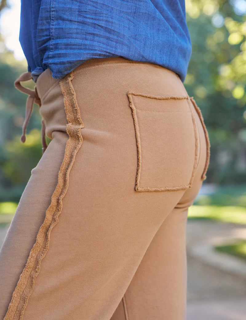 Close-up of a person wearing Frank & Eileen's Bella Sweatpant in tan, featuring 100% cotton fabric, raw edge seams, and a back patch pocket, paired with a tucked-in blue shirt; blurred green outdoor background.